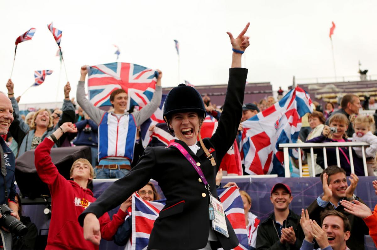 A female Para equestrian athlete poses for a photograph in front of the stands. Spectators are holding the GB flag